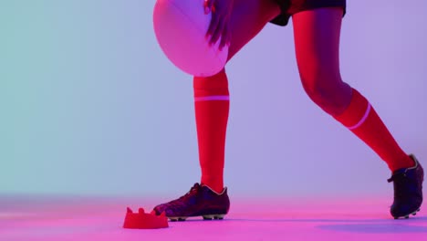 african american female rugby player crouching with rugby ball over neon pink lighting