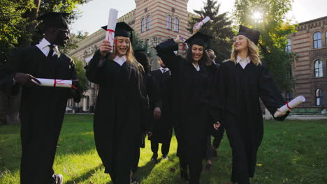 graduados masculinos y femeninos multiétnicos con vestidos y gorras tradicionales negros caminando alegremente y hablando con sus diplomas en las manos del edificio de la universidad