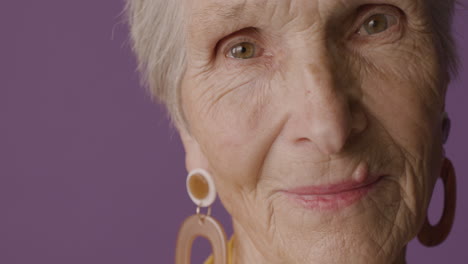 close up view of senior woman with short hair and green eyes wearing earrings posing and smiling at camera on purple background