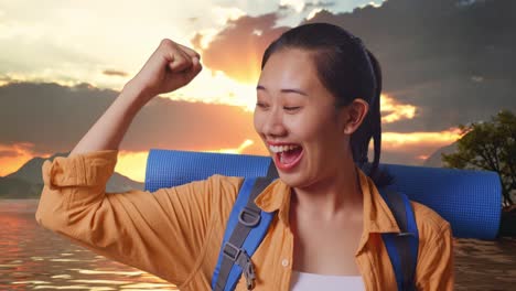 close up de una excursionista asiática con mochila de montañismo sonriendo y flexionando su bíceps mientras está de pie en un lago durante el atardecer