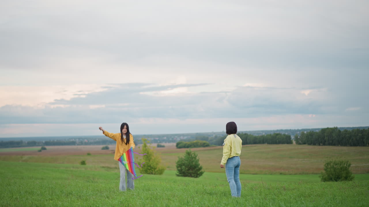 Dois amigos soltando pipa em uma colina com vista para o campo aberto sob um céu nublado, usando jaquetas e jeans coloridos, o vento elevando a pipa para o alto, horizonte tranquilo se estendendo à distância, luz suave da noite