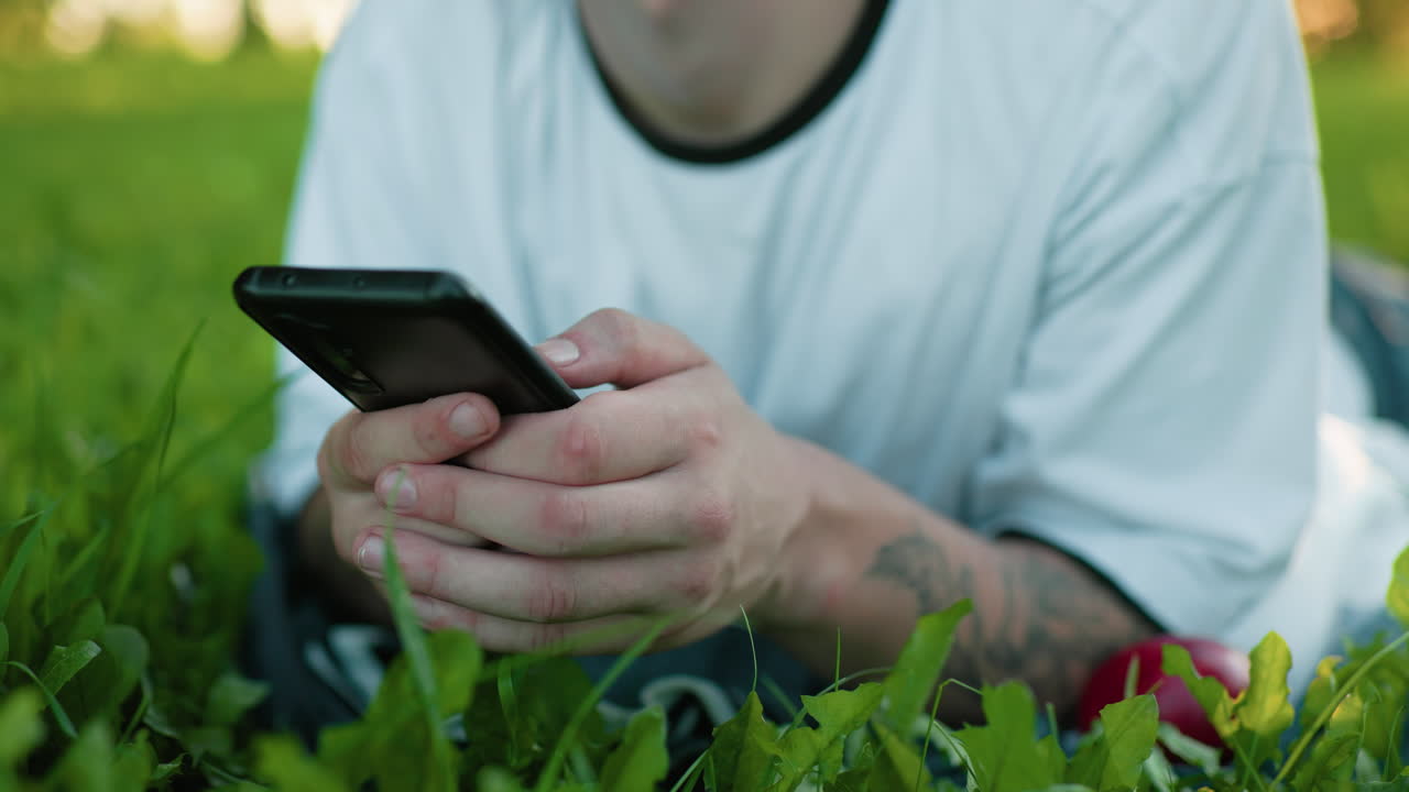 primer plano de un hombre tendido en un campo de hierba usando un teléfono inteligente con una mano tatuada visible, mostrando un momento pacífico al aire libre rodeado de exuberante vegetación