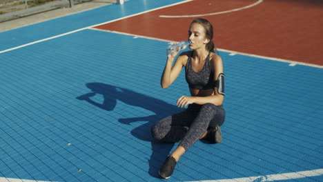 mujer fitness sentada en la cancha deportiva y bebiendo agua fría después del entrenamiento en un día de verano