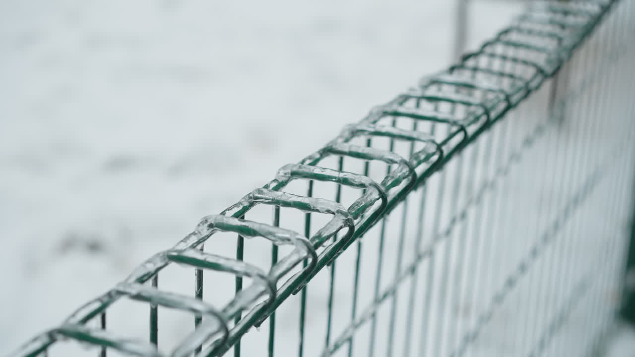 primer plano de una valla metálica verde cubierta de hielo, que muestra hielos colgando de los cables contra un fondo borroso y nevado del parque, enfatizando el marcado contraste entre las texturas congeladas