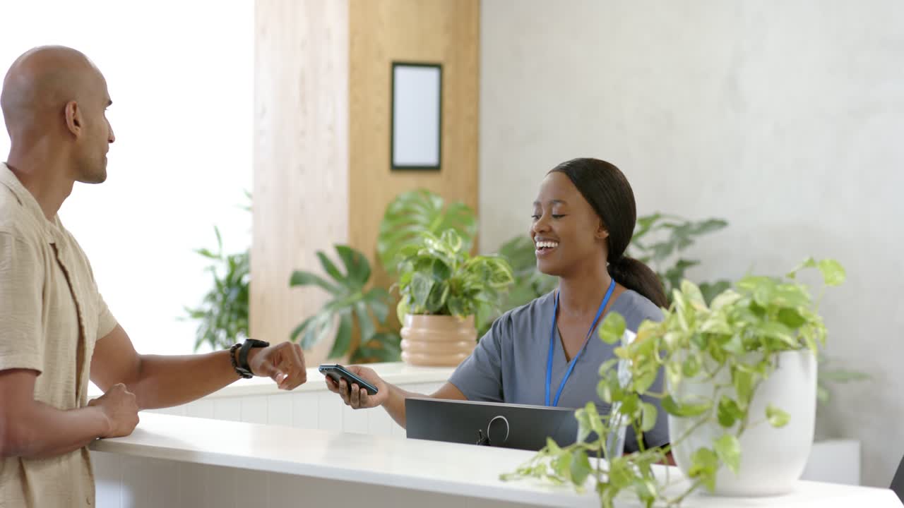 Uomo adulto che controlla il polso, toccando l'orologio alla scrivania; receptionist afroamericana in divisa medica che usa un lettore.