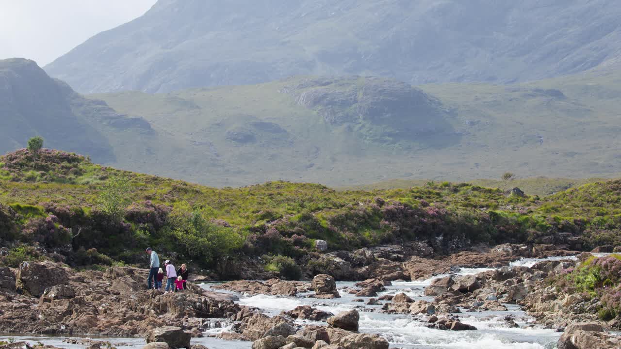 Due persone camminano accanto a un fiume in pieno giorno, circondate da montagne e terreni impervi