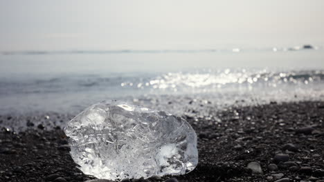 Trozo-De-Iceberg-En-Una-Playa-De-Arena-Negra,-Un-Océano-Brillante-En-El-Fondo,-De-Cerca