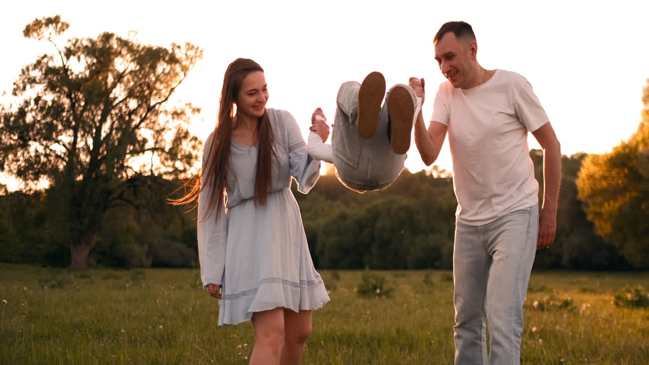 엄마와 아빠는 아이를 흔들고 손에 놀고 있습니다. 소년은 해가 지는 여름에 길거리에서 부모와 함께 놀고 행복합니다.