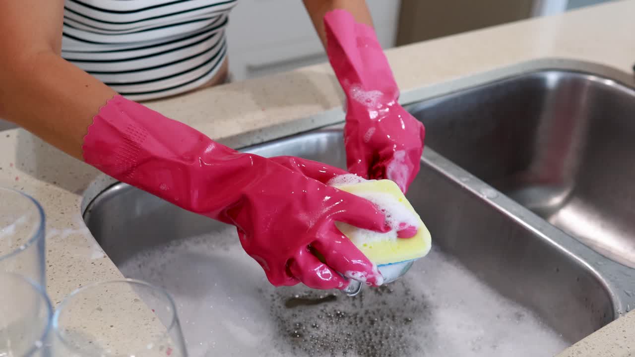 A person cleans a colander in a kitchen sink using pink gloves and a sponge