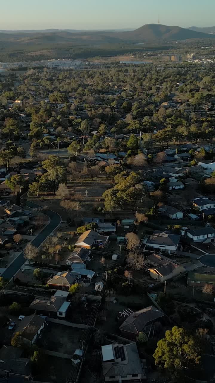 Un dron vertical vuela sobre casas residenciales en los suburbios de Rivett y Western Creek al sur de Canberra. La torre Black Mountain se ve a la distancia, con árboles y vegetación entre las casas al final de la tarde.