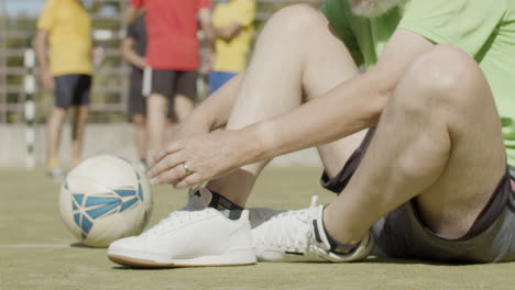 sportive senior football player sitting on pitch and lacing sneakers