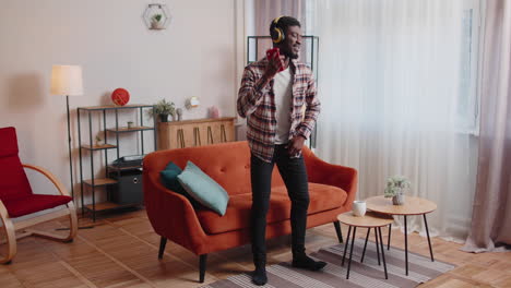 african american young man in headphones listening music dancing, singing in living room at home