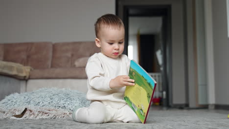 curious little boy sitting on the carpet looking through a book. kid puts a book on the floor and approaches camera.