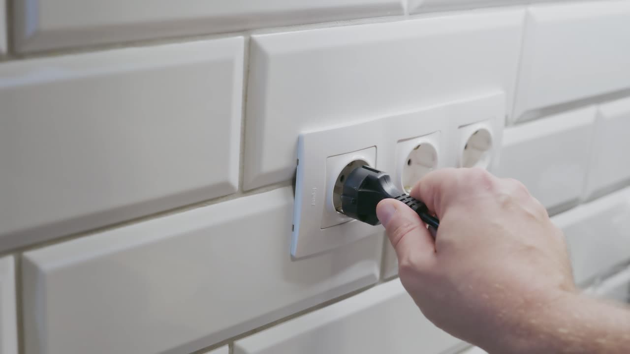 A close-up of a person’s hand inserting a black power plug into a white electrical socket on a tiled wall. The video illustrates a common household activity related to electricity and safety