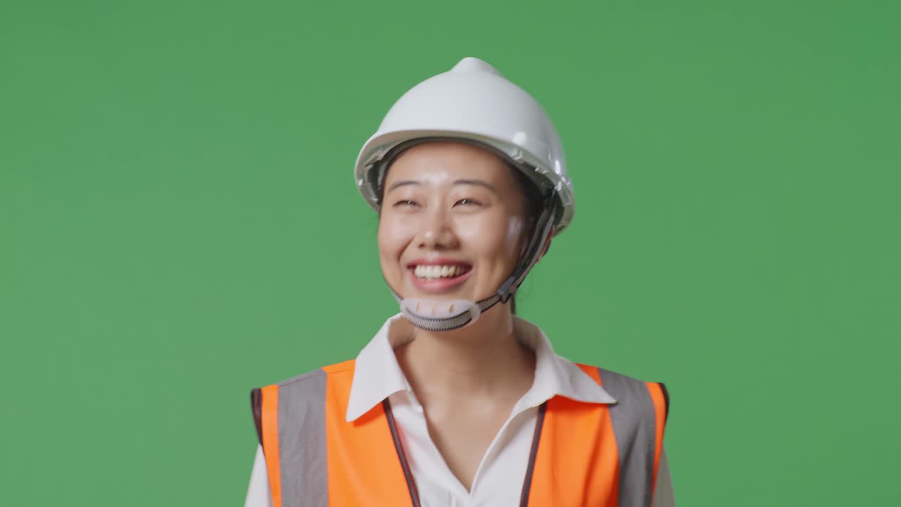 안전 헬을 입은 아시아 여성 엔지니어가 녹색 스크린 배경 스튜디오에서 걸어가는 동안 손을 흔들고 있습니다.