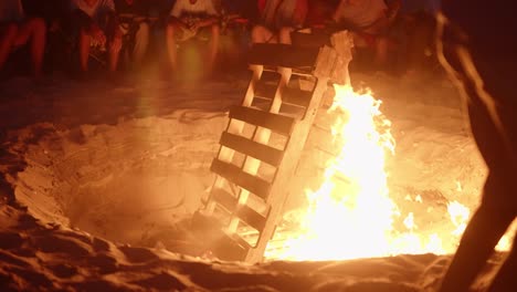 hoguera gigante en un campamento en la playa, llamas encendiéndose a cámara lenta y quemando paletas de madera mientras los campistas miran