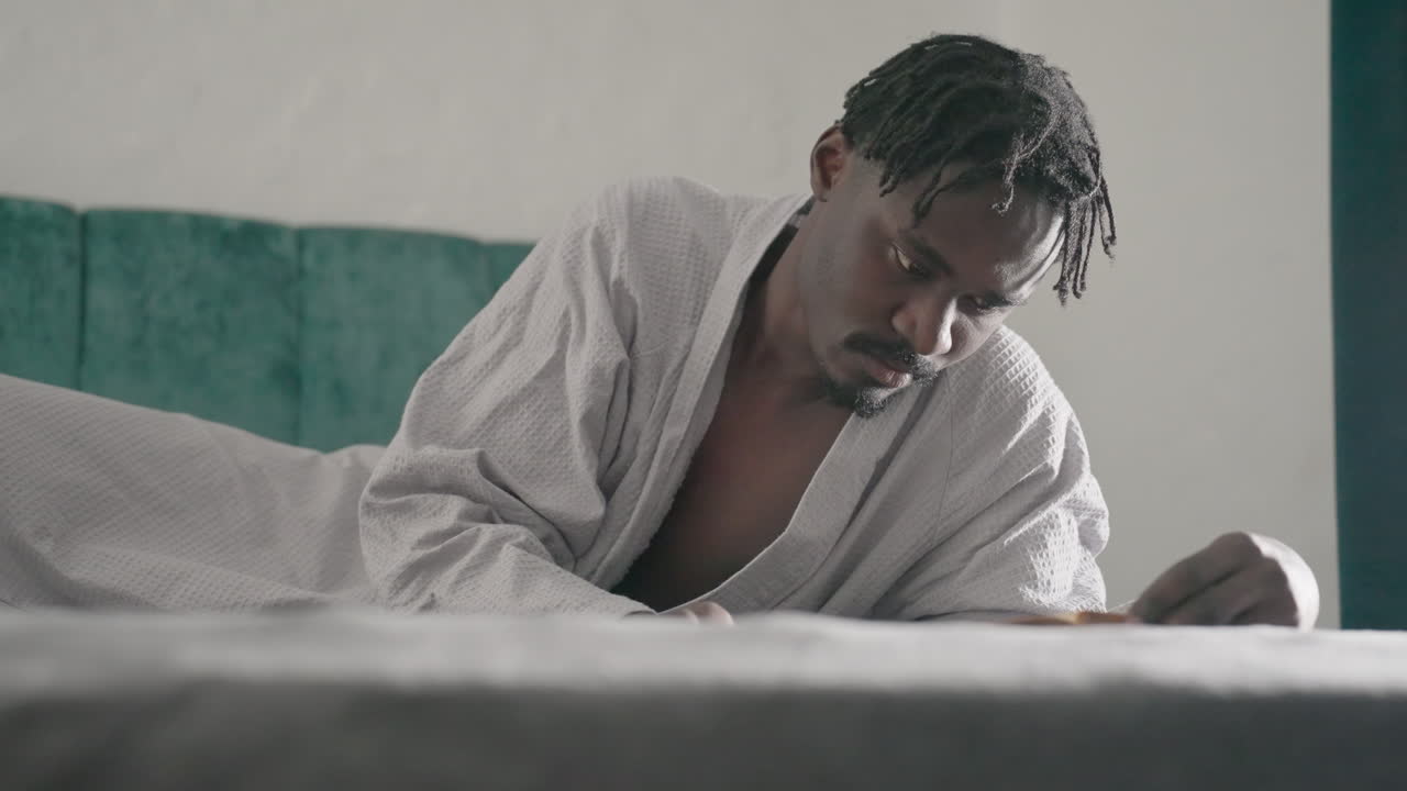 Man in white robe lying on bed, intently reading large book, focused on content. Soft natural light from window illuminates peaceful indoor space, creating serene atmosphere for deep reflection