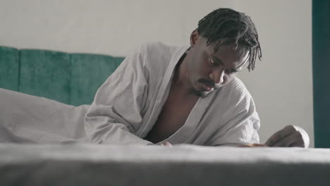 man in white robe lying on bed, intently reading large book, focused on content. soft natural light from window illuminates peaceful indoor space, creating serene atmosphere for deep reflection
