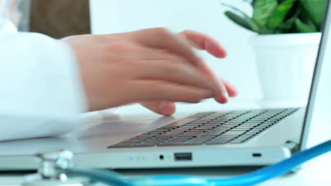 el concepto de medicina, el médico trabaja, las manos de un hombre con una túnica blanca imprimen el texto en la computadora portátil, en la mesa blanca un fanendoscopio y una planta verde dormida