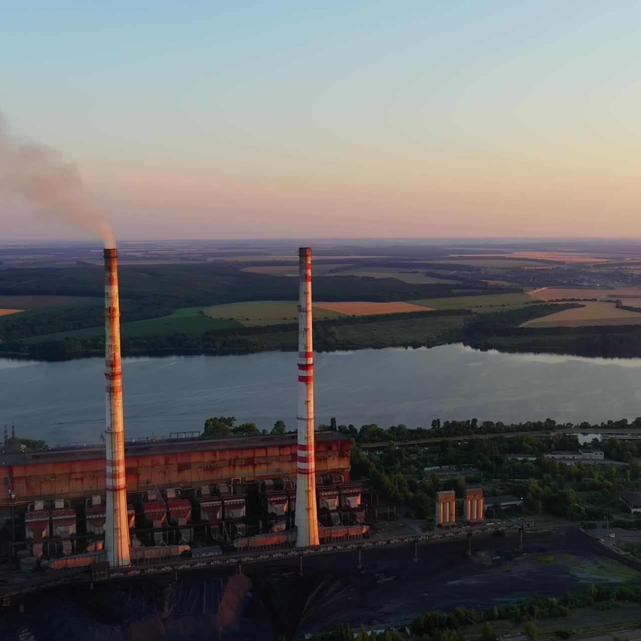 Chemical plant among beautiful nature. Industrial factory with smoking pipe near the river at sunset. Pipe emits harmful emissions and pollute the environment. Aerial view.