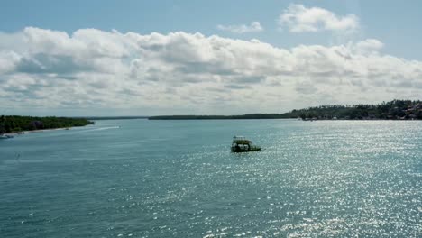 Right-trucking-aerial-drone-shot-following-a-small-transport-boat-sailing-on-a-tropical-turquoise-river-from-the-Restinga-beach-to-the-Barra-do-Cunhaú-beach-in-Rio-Grande-do-Norte,-Brazil