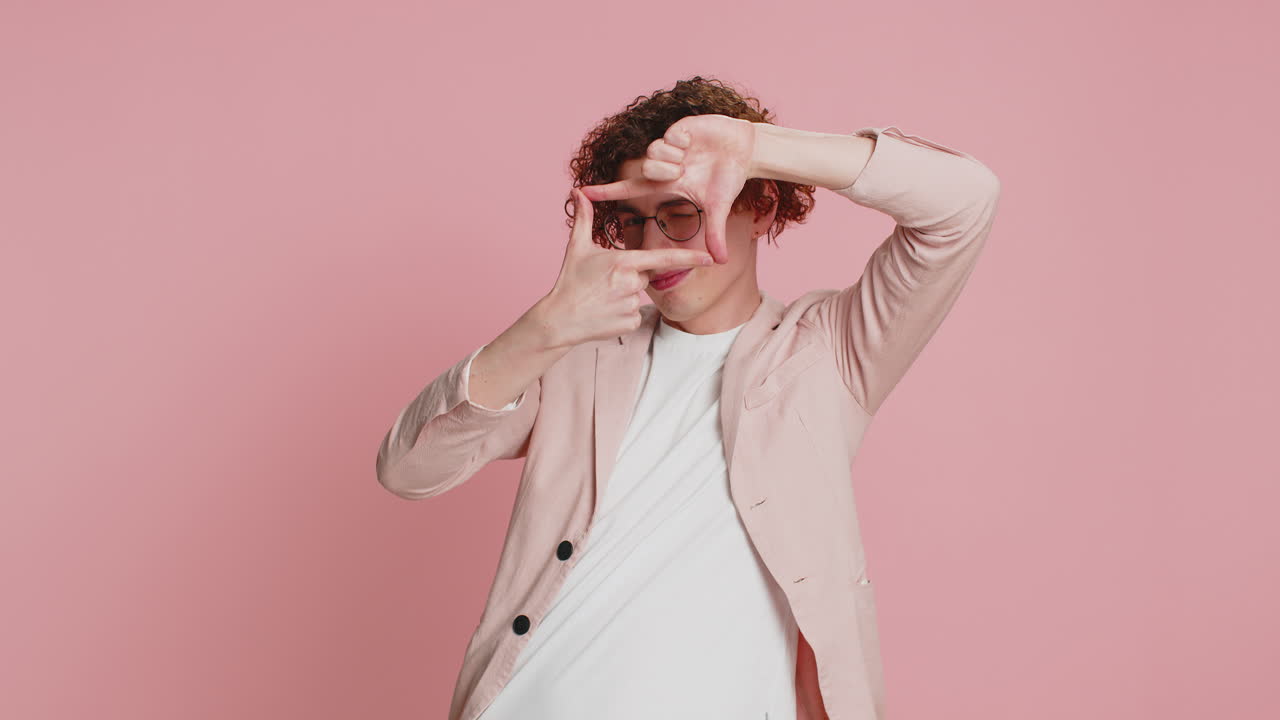 un joven con una chaqueta rosa y gafas sonríe y enmarca la cámara con sus manos