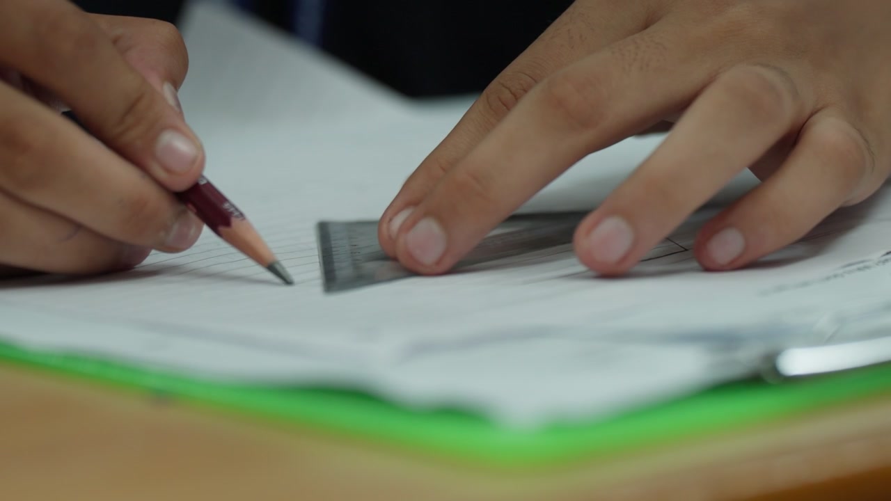 Close-up of hands using a triangular scale ruler and pencil to draw straight lines on white paper. The student works methodically on technical drawing with green folder visible beneath the paper.