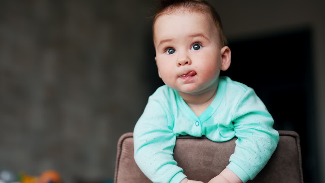 Serious interested kid standing on the chair looks around. Curious baby studying surrounding leaning on a chair back. Low angle view.