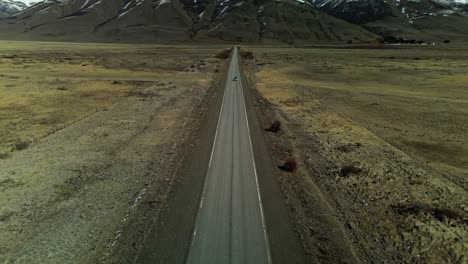 Toma-Aérea-Inclinada-Hacia-Arriba-Que-Muestra-El-Auto-En-La-Carretera-En-La-Patagonia-Con-Cordillera-Y-Picos-Nevados-En-El-Fondo,-Argentina