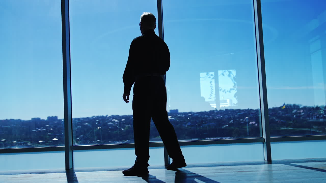 Full-length size portrait of an adult male wearing black official clothes. Man comes up to the panoramic window and looks at sunny cityscape. Low angle view.
