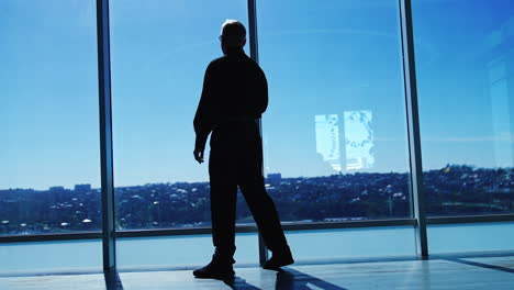 full-length size portrait of an adult male wearing black official clothes. man comes up to the panoramic window and looks at sunny cityscape. low angle view.