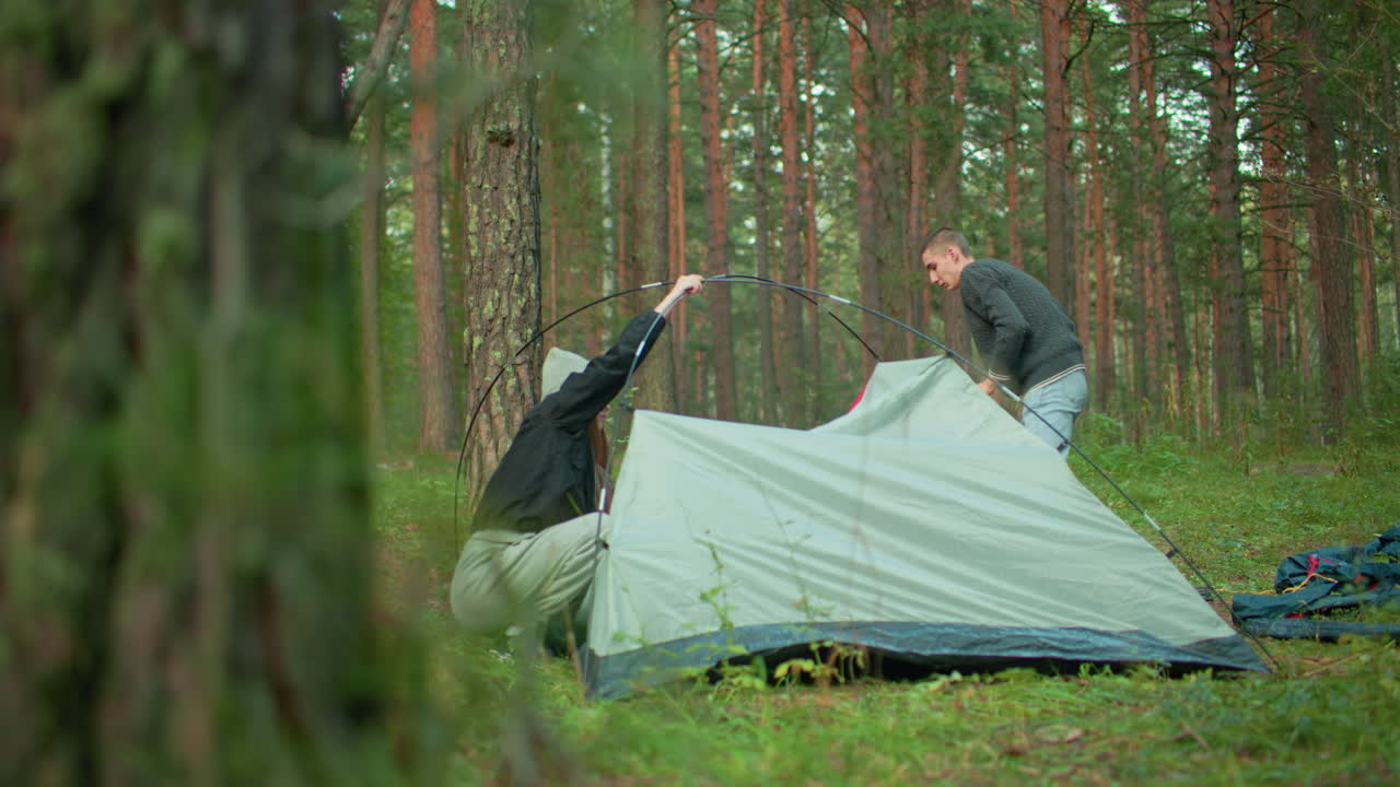 man and woman work together to assemble camping tent in lush forest as one lifts flexible pole and other secures fabric on grassy ground surrounded by tall trees