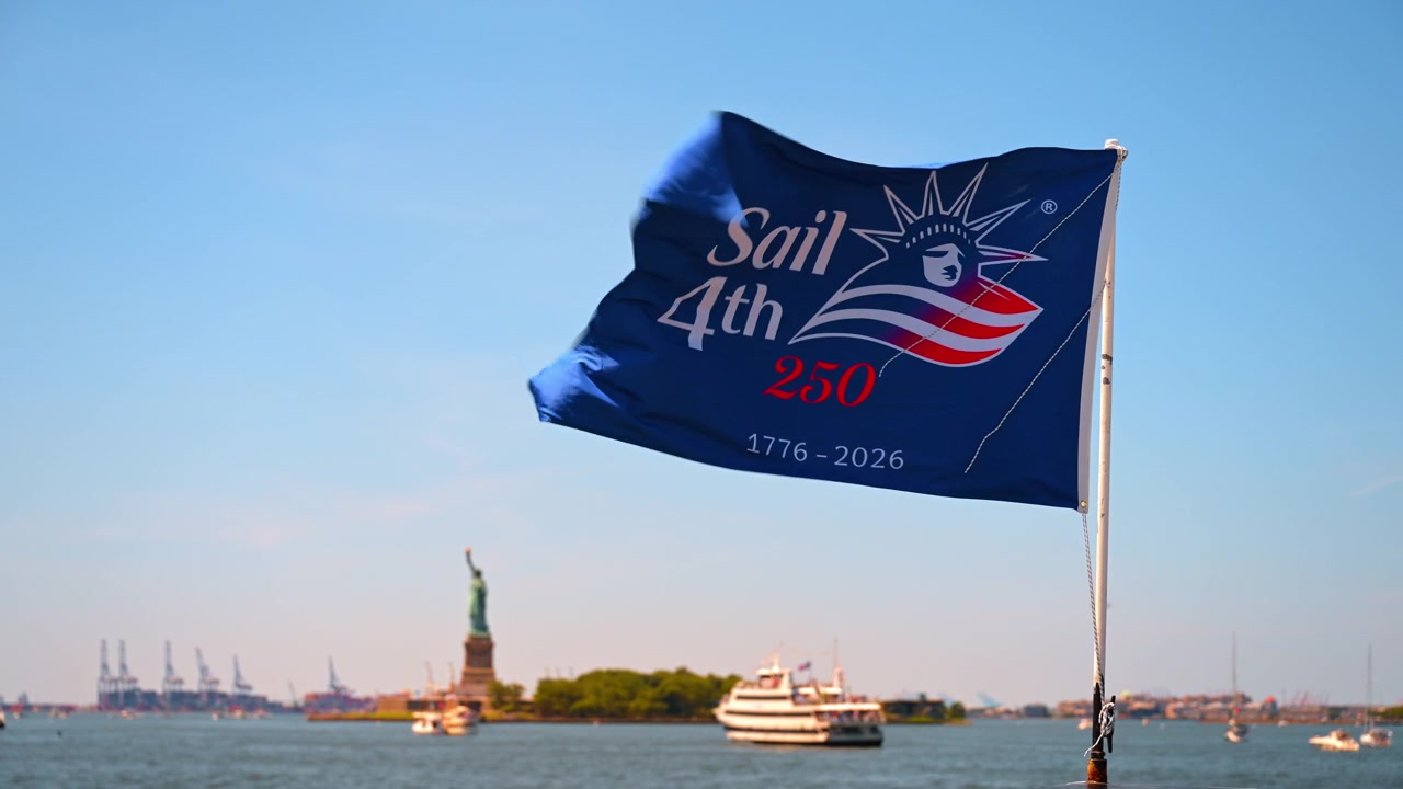 Bandiera Sail4th 250 sopra la Statua della Libertà. Uno striscione blu dell'evento sventola sul porto di New York mentre le barche passeggeri attraversano l'acqua illuminata dal sole