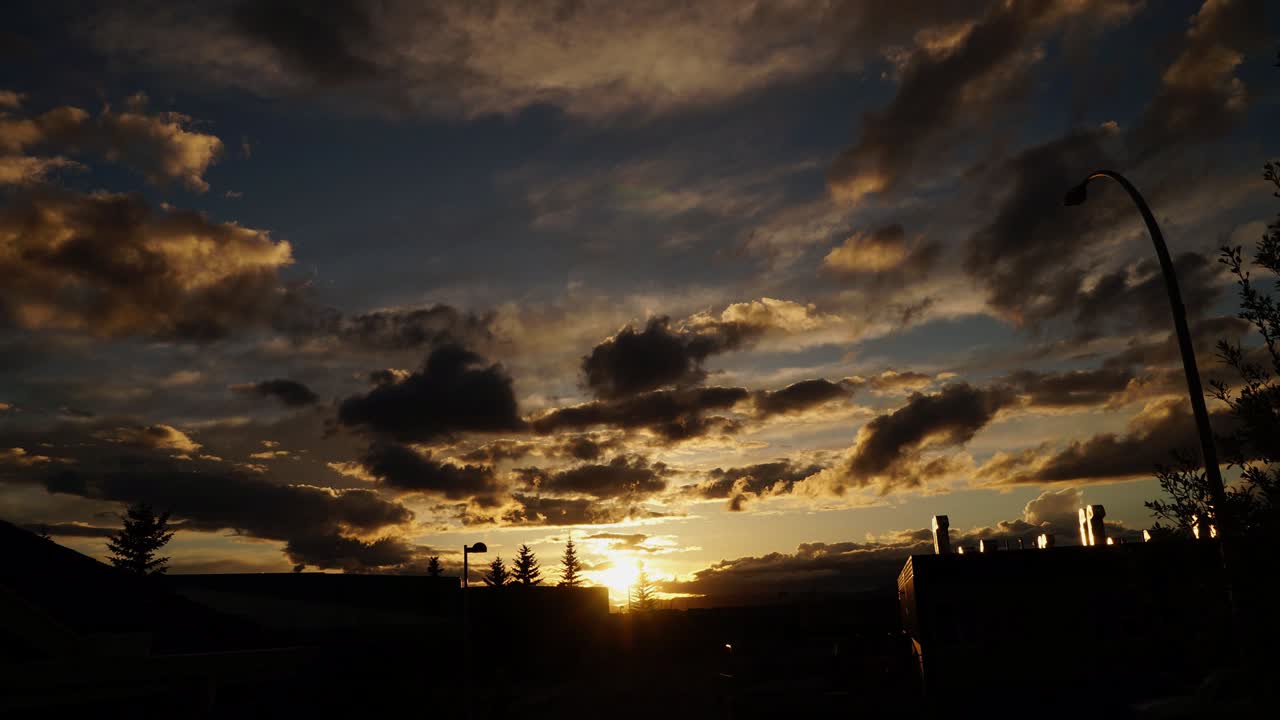 Zeitrafferaufnahme eines Sonnenuntergangs mit Gebäudesilhouetten, während im Sommer mehrere Wolkenschichten in zwei Richtungen vorbeiziehen. Alberta, Kanada