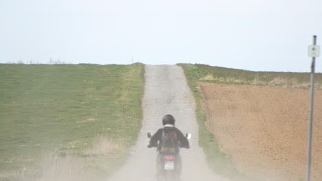 Ein-Motorradfahrer,-Der-Auf-Einer-Staubigen-Schotterstraße-Zwischen-Zwei-Wiesen-In-Deutschland-Fährt