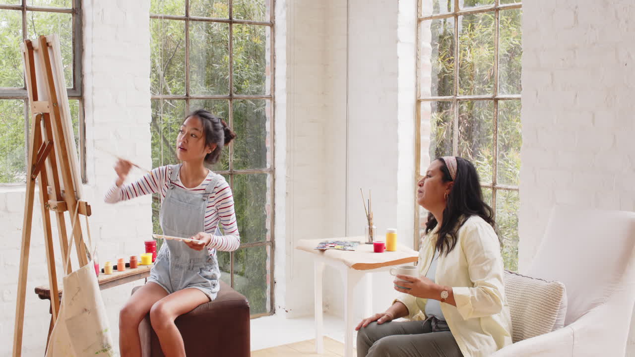 Multiracial young woman painting on canvas with grandmother enjoying coffee, at home