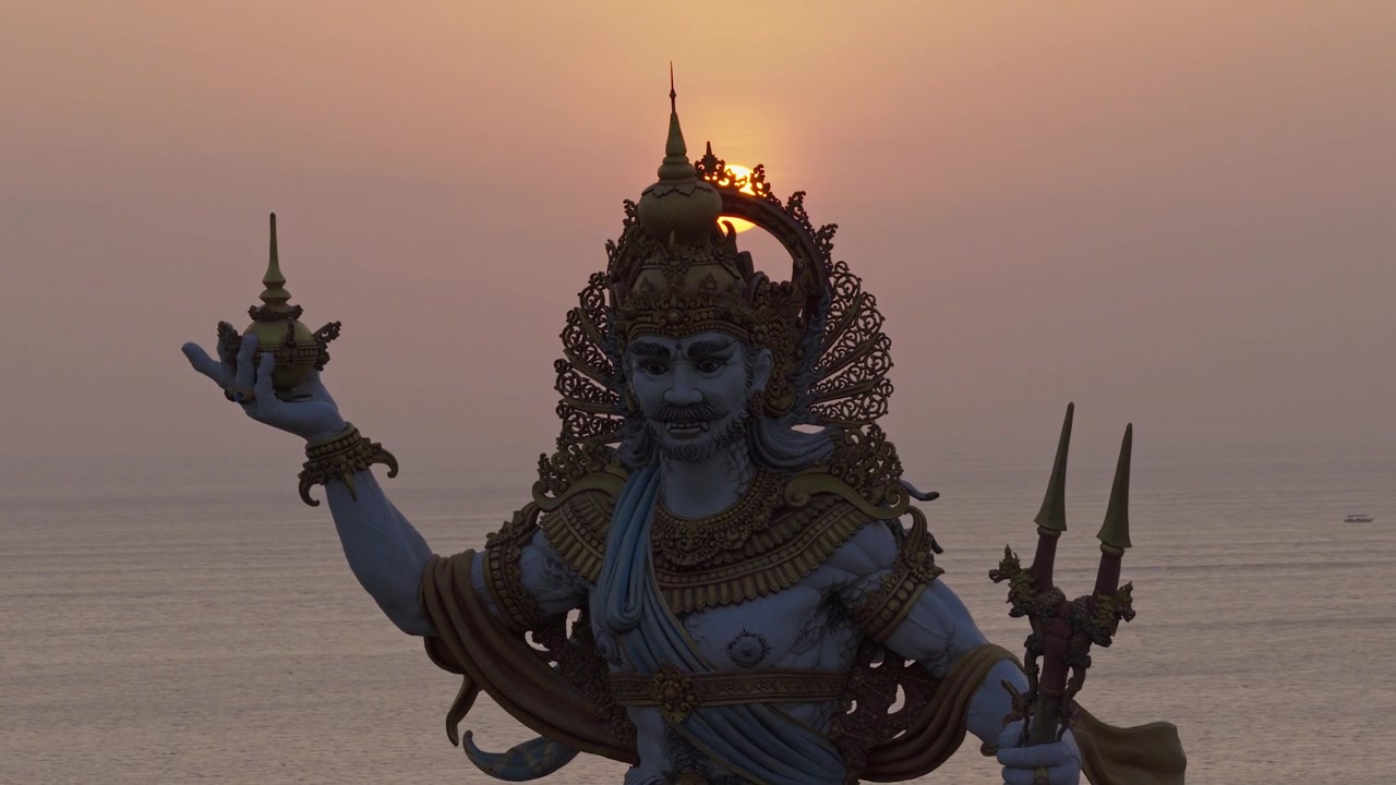 Tropical sunset over Kuta Beach with a monumental Hindu statue overlooking the sea showcasing intricate Balinese artwork traditional ornaments and a hazy golden horizon orbit aerial drone footage