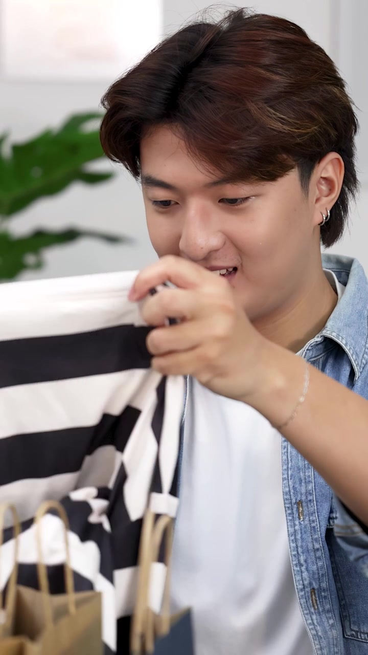 Un giovane uomo asiatico sorridente scarta una camicia a righe da un sacchetto di carta in un luminoso salotto.