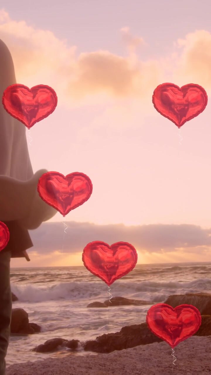 Molti palloncini a forma di cuore fluttuanti, su sfondo di una giovane coppia che si abbraccia in riva al mare.