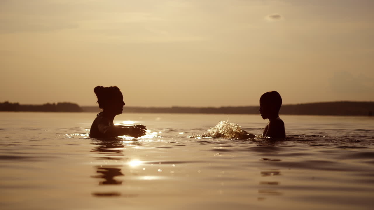 Silhouette of a woman and a boy in water at sunset. Mother and son are splashing water at each other in the river in the evening.