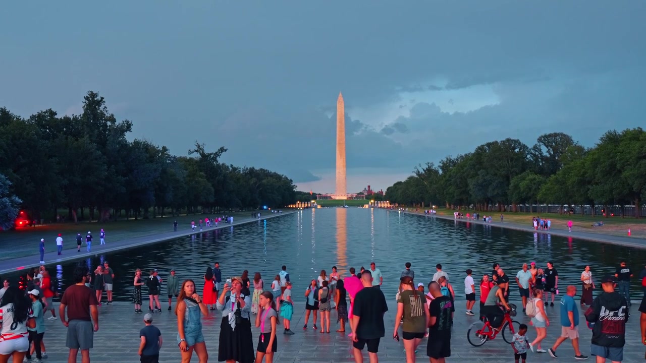 Washington, USA, 12 giugno 2026: Folla che si raduna presso il Reflecting Pool con il Washington Monument, Washington, D.C. Turisti lungo il bordo dell'acqua con il riflesso dell'obelisco sotto un cielo crepuscolare blu nuvoloso.