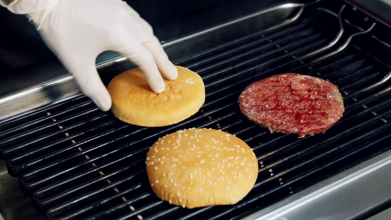 Roasting burger components on hot grill. Chef's hand in glove turning up halves of buns during frying in kitchen. Slow motion