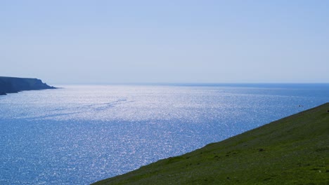 beautiful view across the sea with sun reflecting and glistening on calm water and clear blue sky as cliff coastline comes into view. filmed on gower pensinsula, wales uk.