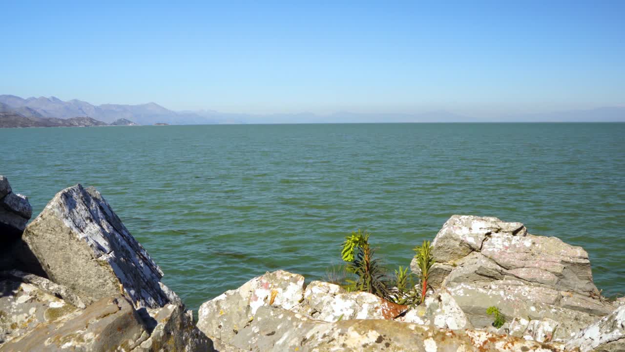 Beautiful white rocks and green herbs on shore of lake surrounded by mountains on a sunny winter day