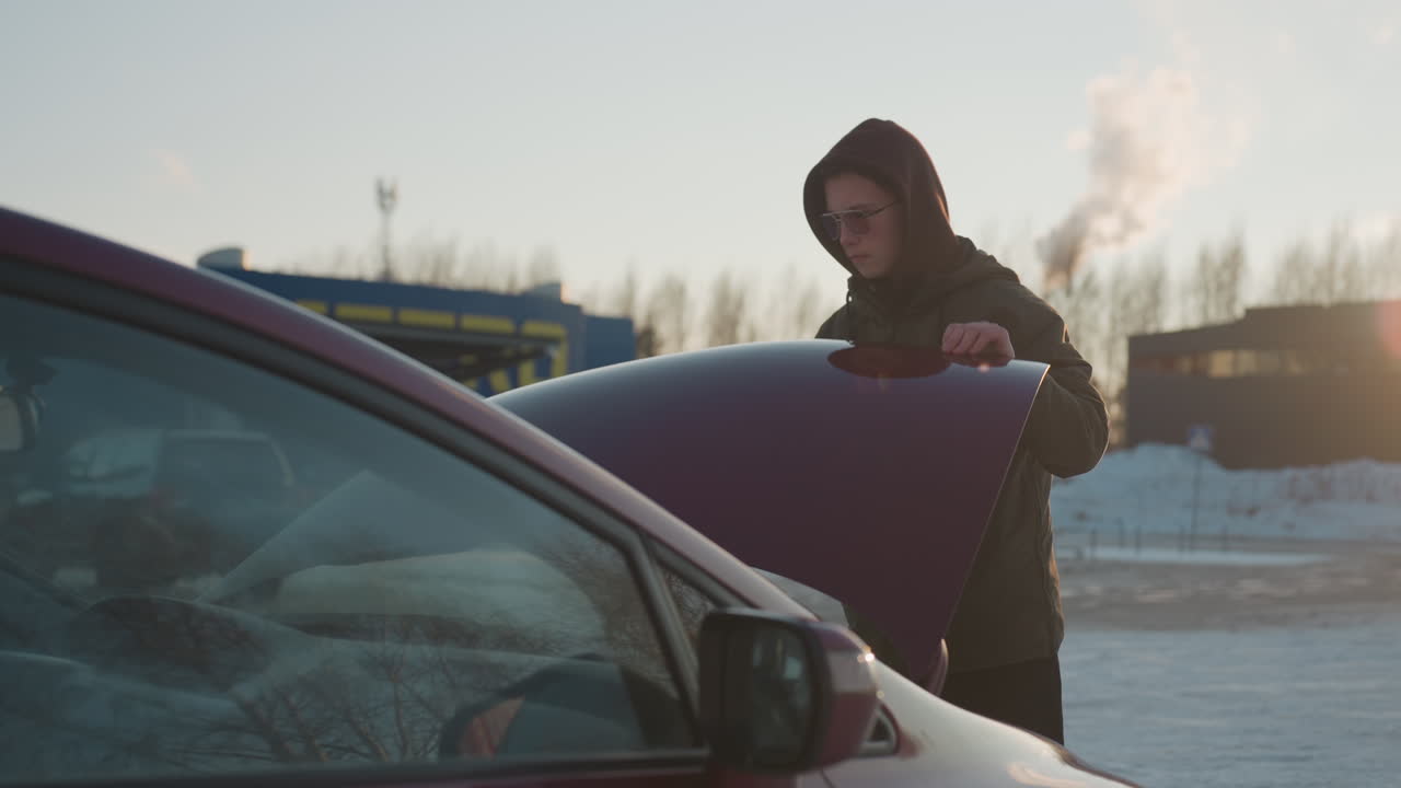 Man wearing hoodie closes raised car bonnet with visible tension in arm as winter sunlight illuminates background with frosted parking area and bare trees in cold outdoor setting