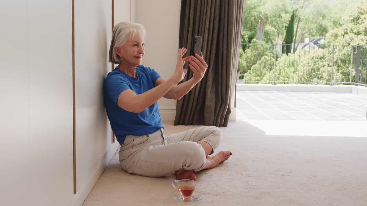 Senior woman video chatting on smartphone, sitting on floor with tea, at home