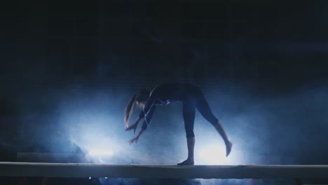 the girl is a professional athlete performs gymnastic acrobatic trick on a beam in backlight and slow motion in sports gymnastic clothing. smoke and blue. jump and spin on the balance beam