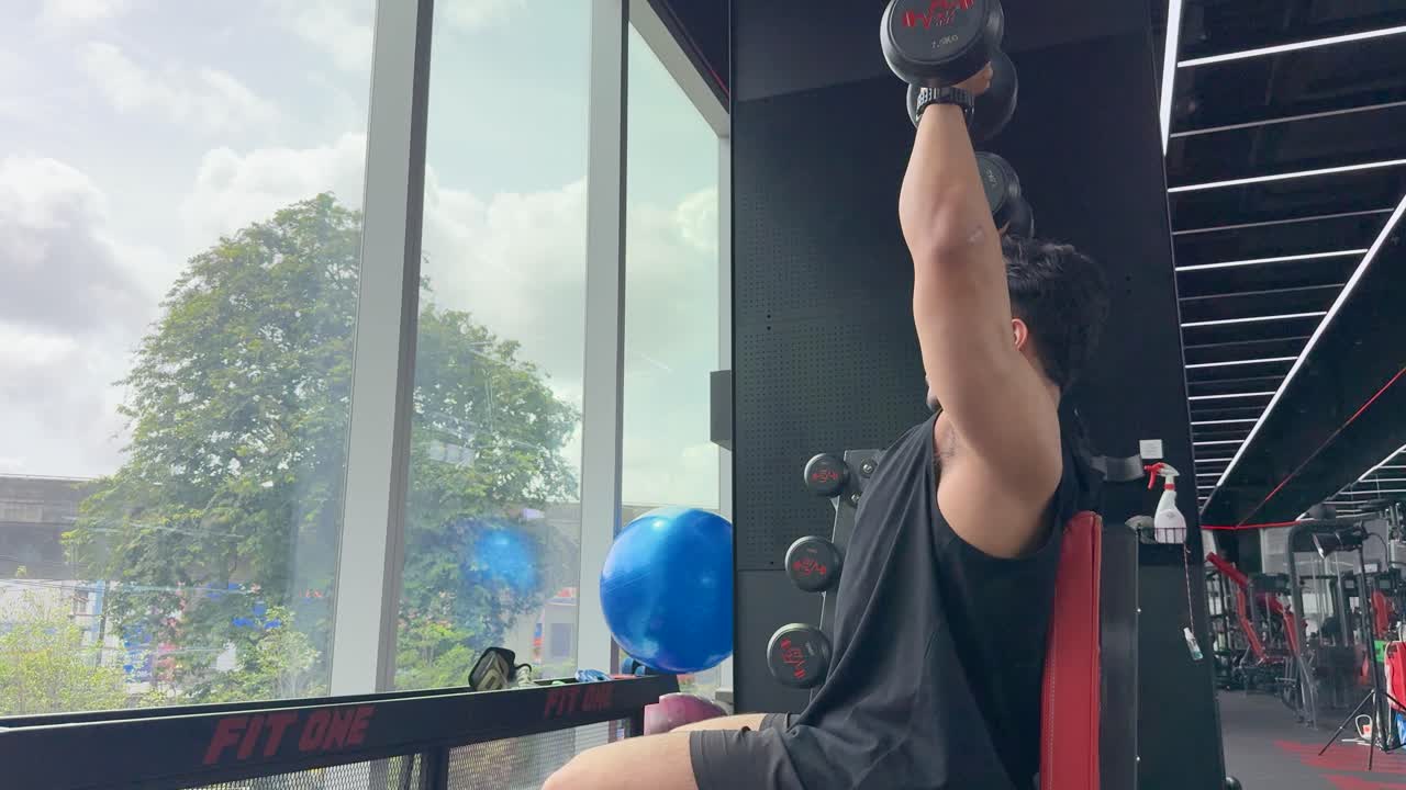A person performs bicep curls in a gym, seated by a large window with natural light