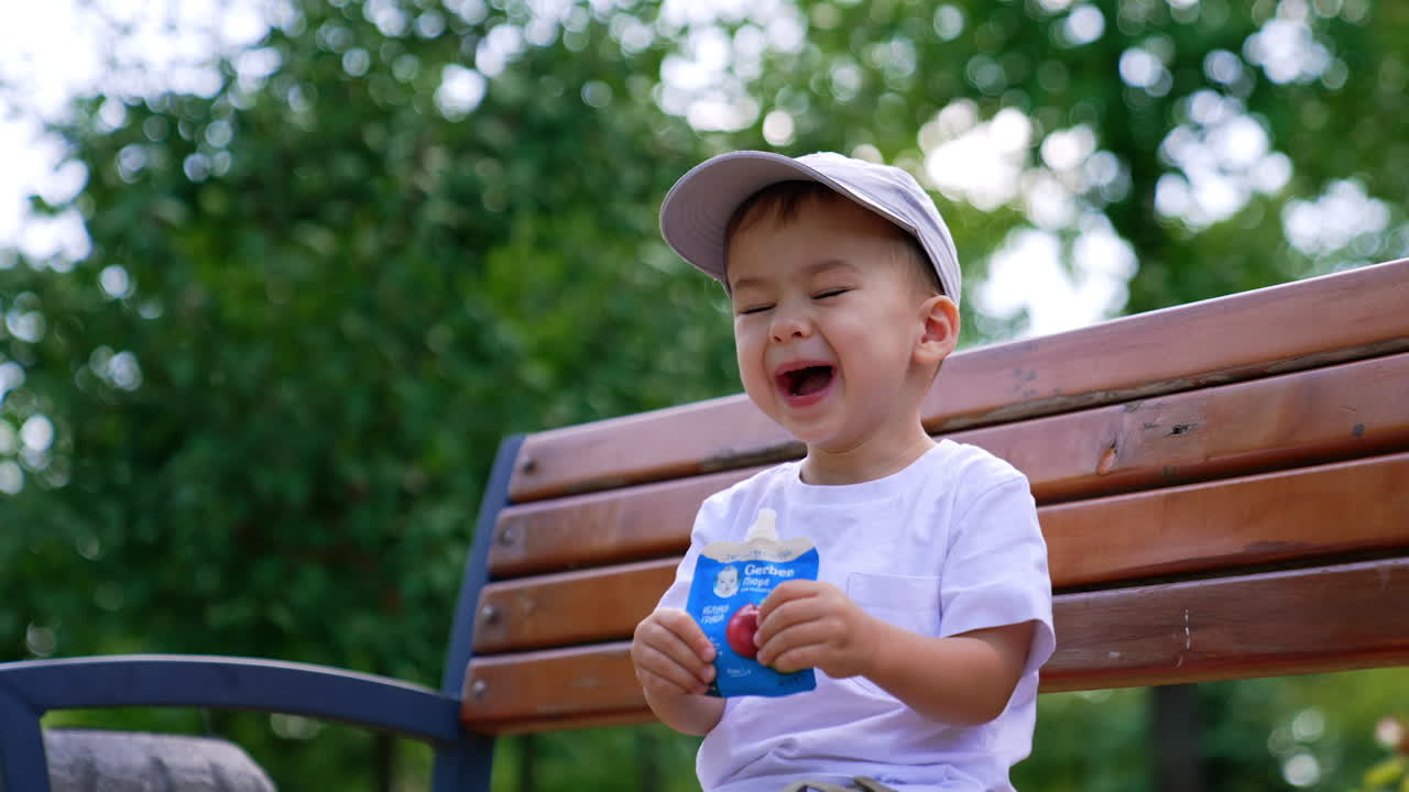 Lovely baby boy in cap sits on the bench drinking the juice. Caucasian toddler having a healthy snack outdoors. Low angle view.