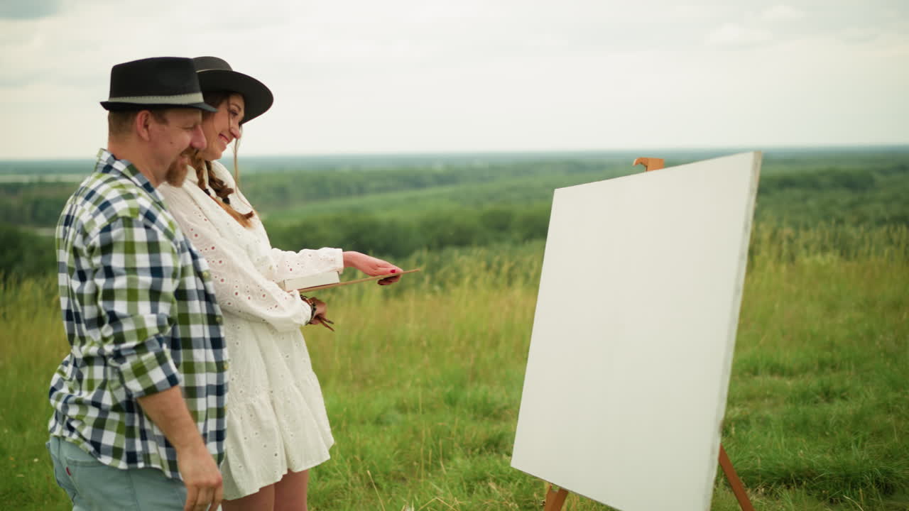 Couple stands in a grassy field, preparing to paint. The man, in a black hat and plaid shirt, and the woman in a white dress share a moment of laughter as they look at a large white board on an easel